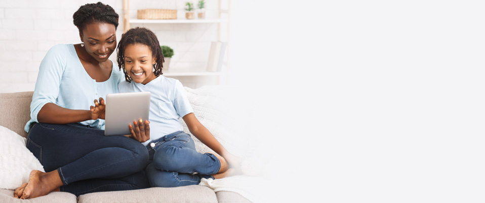 A woman and girl site on the couch interacting with a tablet device.